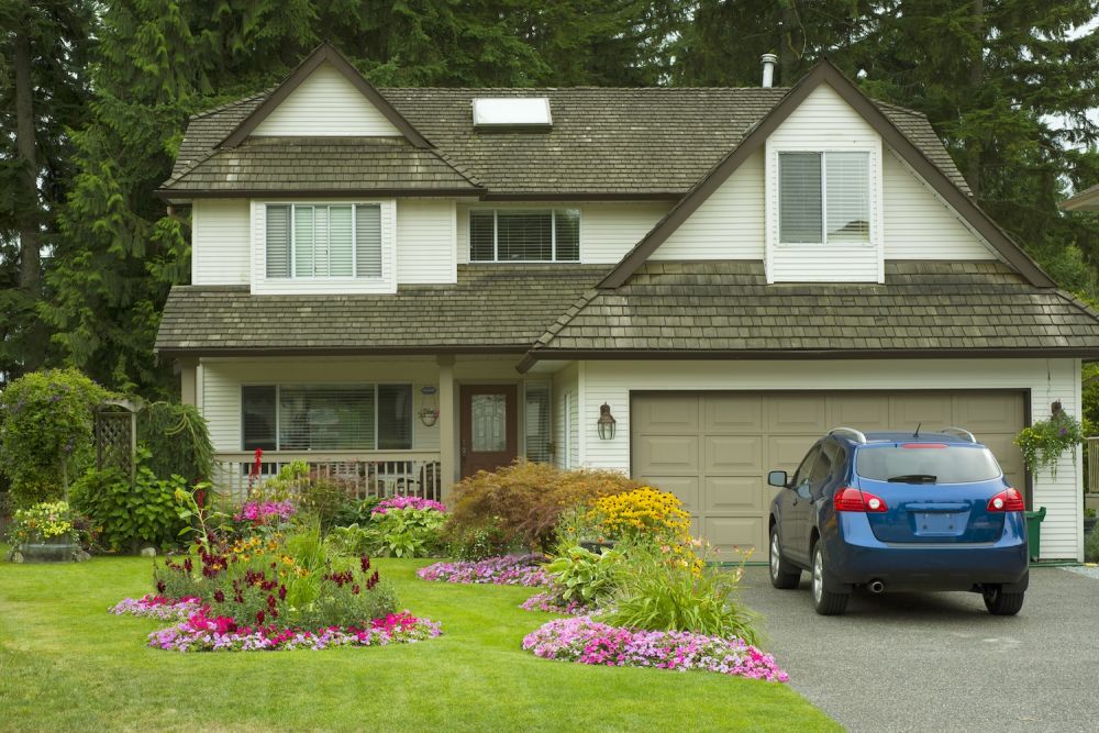 Charming suburban home with vibrant garden and parked blue car in driveway, surrounded by lush green trees.