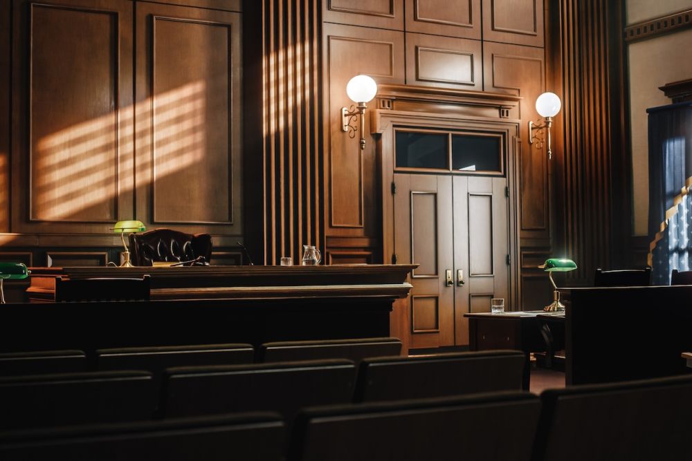 Elegant courtroom interior with wood paneling, empty seats, judge's bench, and warm sunlight streaming through windows.